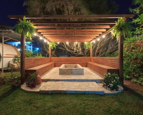 Illuminated wooden gazebo with fireplace and benches in garden setting at night.