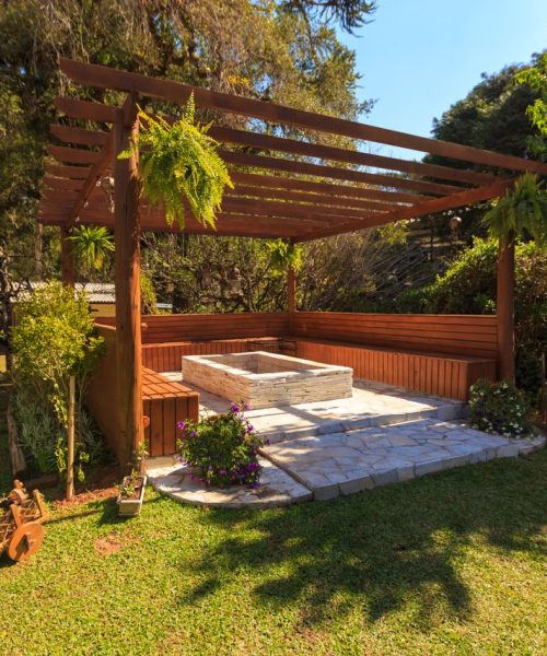 Spacious wooden gazebo in a lush garden setting with green foliage and stone flooring.
